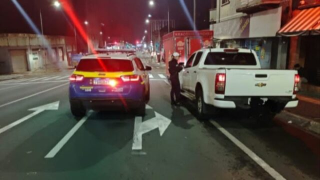 Viatura oficial e caminhonete branca parada em via pública durante abordagem policial à noite