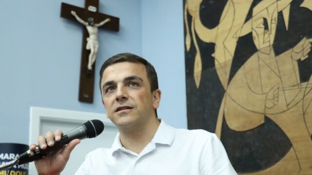 Deputado federal Aliel Machado segura microfone e discursa em plenário com crucifixo e painel de madeira ao fundo