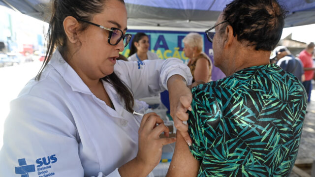 Profissional de saúde com jaleco do SUS aplicando vacina no braço de um idoso em Ponta Grossa