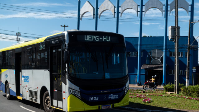 Ônibus urbano cinza, amarelo e azul com letreiro digital indicando UEPG-HU rodando em rua de Ponta Grossa
