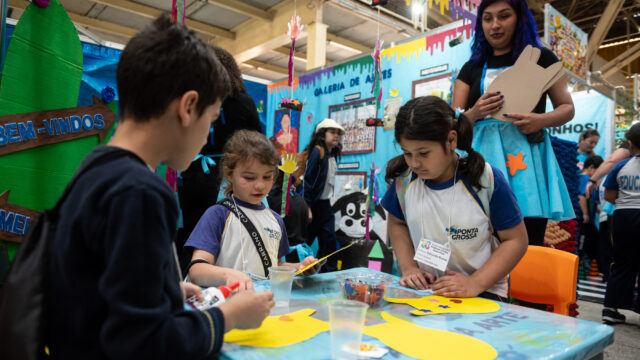 Crianças sentadas à mesa decorada fazendo artesanato com papel em um evento educacional em Ponta Grossa