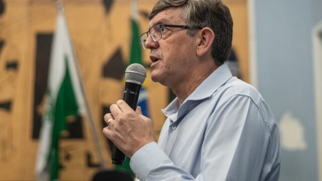 Homem de óculos e camisa social azul clara segura microfone preto durante pronunciamento