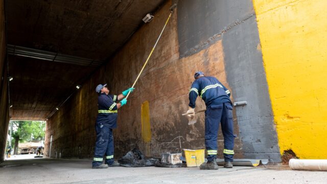 Dois operários pintam as paredes de concreto de um túnel com rolos presos a cabos longos