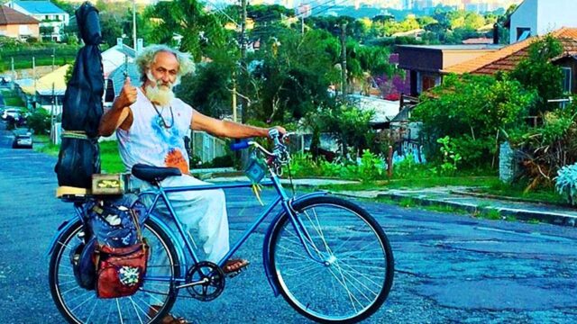 O músico Plá com barba branca e cabelos longos posa sorrindo ao lado de sua bicicleta azul em rua arborizada