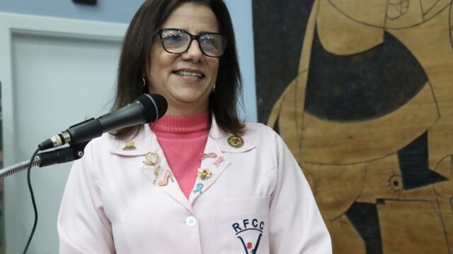 Mulher com óculos e jaleco rosa com o logotipo da Rede Feminina de Combate ao Câncer discursando no microfone