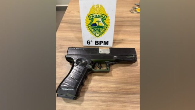 Arma de brinquedo preta e verde sobre uma mesa de madeira junto a um expositor do 6º BPM
