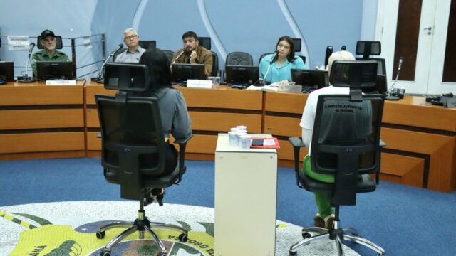 Pessoas sentadas em cadeiras de escritório de costas para a câmera diante da mesa de vereadores em plenário