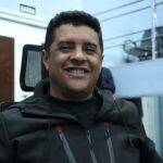 Homem sorrindo veste jaqueta preta em ambiente interno de plenário com porta e placa de saída ao fundo