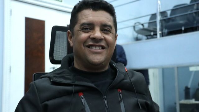Homem sorrindo veste jaqueta preta em ambiente interno de plenário com porta e placa de saída ao fundo