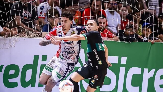 Jogadores de futsal disputam a bola em quadra durante partida eliminatória com torcedores ao fundo