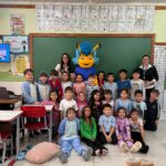 Crianças em sala de aula posam com professores e o mascote azul do programa Ailogs em escola do Paraná