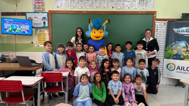 Crianças em sala de aula posam com professores e o mascote azul do programa Ailogs em escola do Paraná