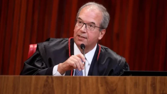 Ministro usando toga preta com detalhes vermelhos em tribunal com painel de madeira ao fundo