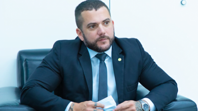 Deputado federal Carlos Jordy sentado em poltrona escura, vestindo terno preto, camisa branca e gravata
