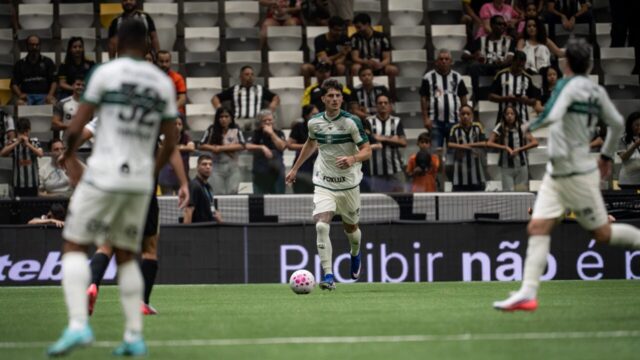 Jogador do Coritiba com uniforme branco e verde conduz a bola em campo diante da torcida na Arena MRV
