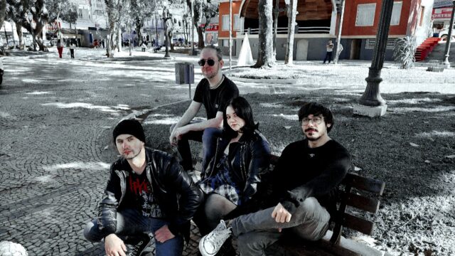 Quatro músicos sentados e agachados em um banco de praça com calçamento de pedras e árvores ao fundo
