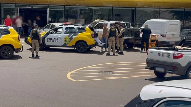 Policiais militares ao lado de viatura e veículos no estacionamento de supermercado em Cascavel