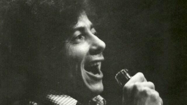 Homem com cabelo cacheado cantando com microfone na mão em apresentação ao vivo