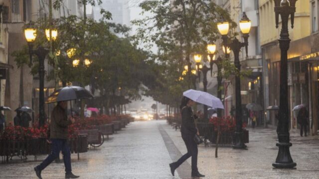 Pedestres com guarda-chuvas caminham por calçadão molhado durante dia chuvoso
