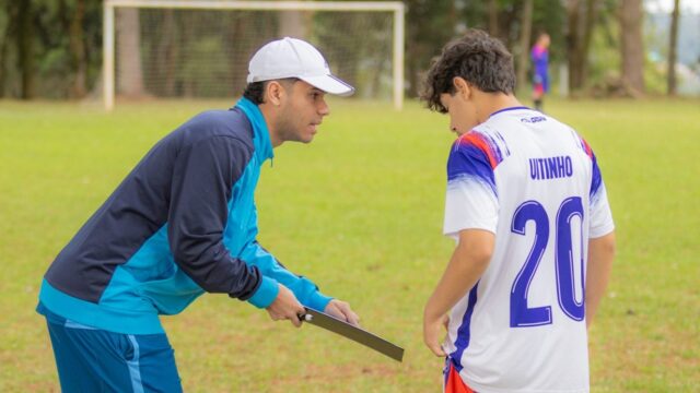Treinador com boné branco conversa com jovem jogador de futebol em campo gramado