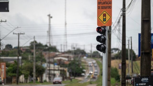 Placa de obras e semáforo recém-instalados à beira de via asfaltada em Ponta Grossa