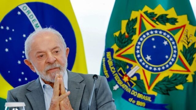 Presidente Luiz Inácio Lula da Silva com as mãos juntas diante das bandeiras do Brasil
