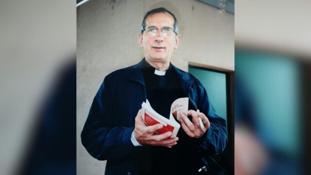 Padre Mário Casassa de óculos e batina escura segurando livro e documento em ambiente interno