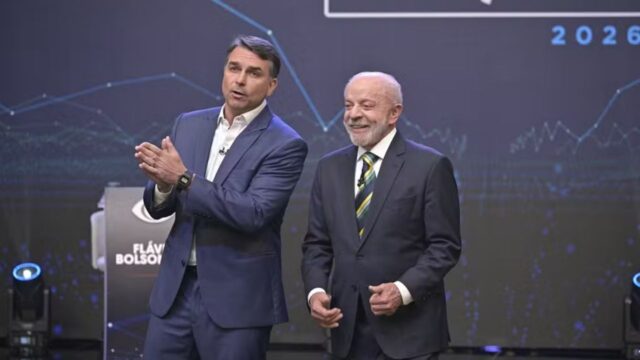 Luiz Inácio Lula da Silva e Flávio Bolsonaro em pé no palco de um debate na TV Bandeirantes