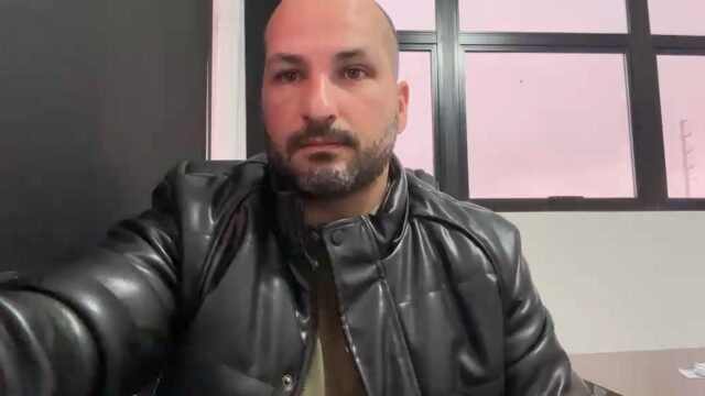 Homem calvo com barba veste jaqueta de couro preta em escritório com janela ao fundo