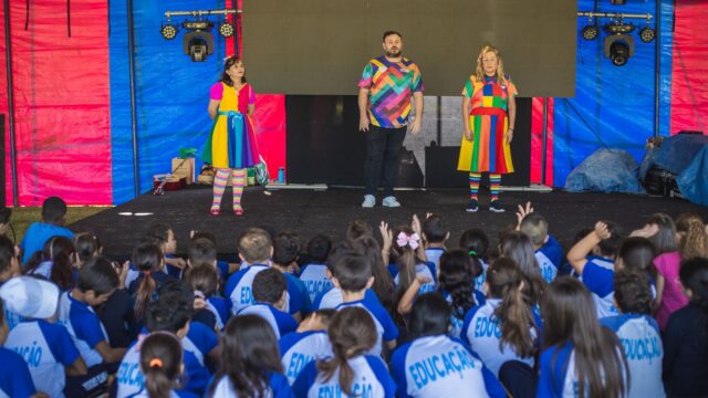Apresentação artística em palco para plateia de crianças uniformizadas com camisas da rede municipal de ensino