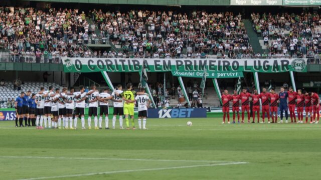 Jogadores de futebol perfilados no gramado de um estádio com torcida nas arquibancadas