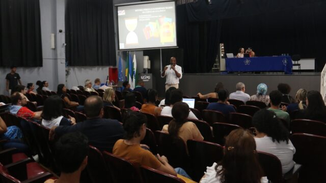 Público assistindo a palestra em auditório com palco e projetor ao fundo