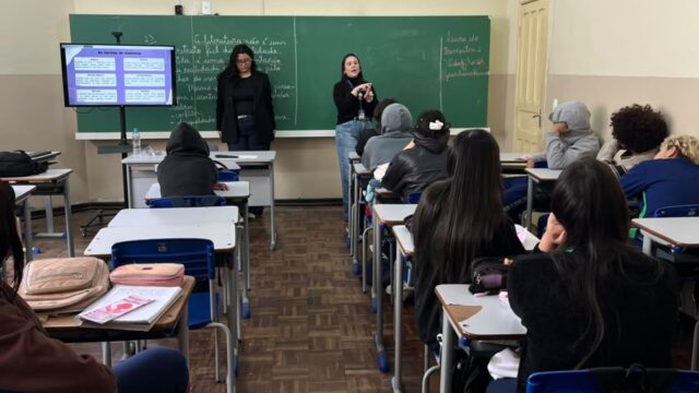 Alunos sentados em carteiras em uma sala de aula assistem a uma palestra sobre violência contra a mulher