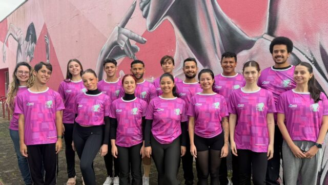 Integrantes da Companhia de Dança de Ponta Grossa com camisas roxas posam para foto em frente a um mural