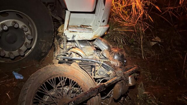 Motocicleta destruída sob a roda de um caminhão em trecho de rodovia à noite