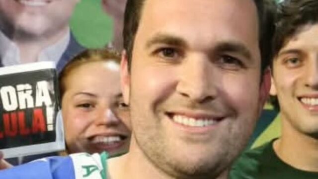 Homem sorridente com camisa verde e bandeira ao fundo, ladeado por apoiadores durante evento político