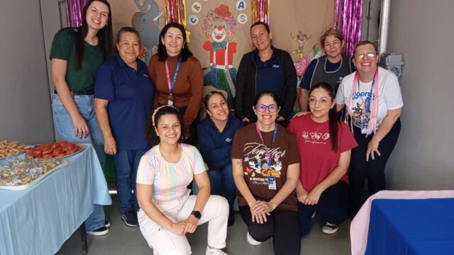 Grupo de dez mulheres trabalhadoras do CRAS Cará-Cará posando sorridentes em frente a um painel decorado para o Dia das Cr…