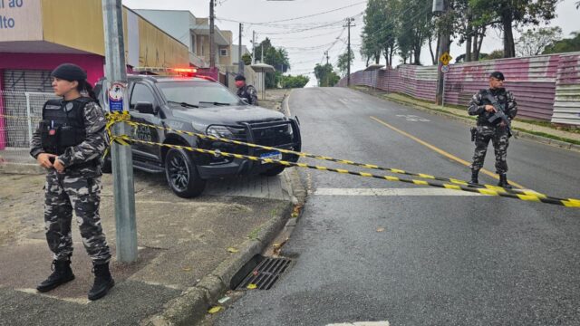 Policiais militares fardados fazem o isolamento com fita amarela em rua com viatura estacionada próximo ao CMEI de Curitiba