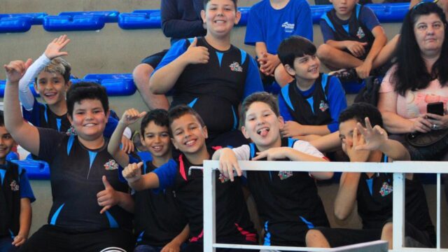 Grupo de estudantes sorri e faz gestos para a câmera nas arquibancadas de um ginásio durante competição escolar