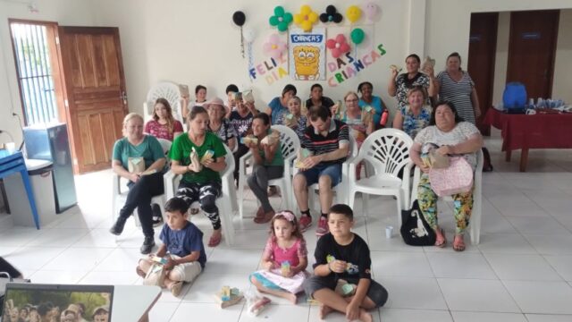 Grupo de crianças e adultos sentados em cadeiras e no chão durante evento com decoração festiva em Ponta Grossa