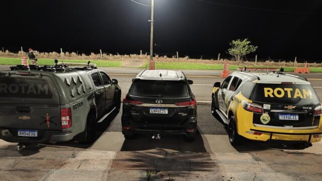 Três veículos estacionados à noite, incluindo viaturas da ROTAM e uma caminhonete preta ao centro