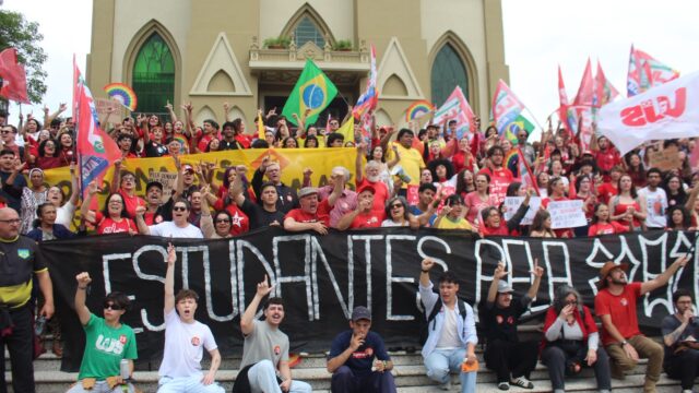 Grupo de manifestantes com bandeiras e uma faixa preta com a inscrição 'Estudantes' sentados e em pé nas escadarias