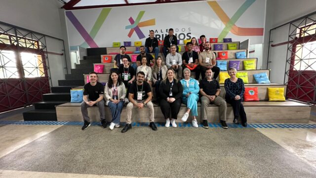 Grupo de pessoas posa sentado em arquibancada com almofadas coloridas durante visita técnica em espaço corporativo