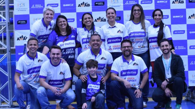 Grupo de dez adultos e uma criança posando sorridentes em quadra de esporte com painel azul ao fundo