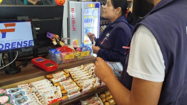 Funcionária da FASPG conversa com comerciante em posto de combustíveis enquanto homem observa prateleira