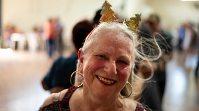 Mulher idosa sorrindo, usando tiara com enfeites natalinos e brincos de argola, em evento do CECON Nova Rússia