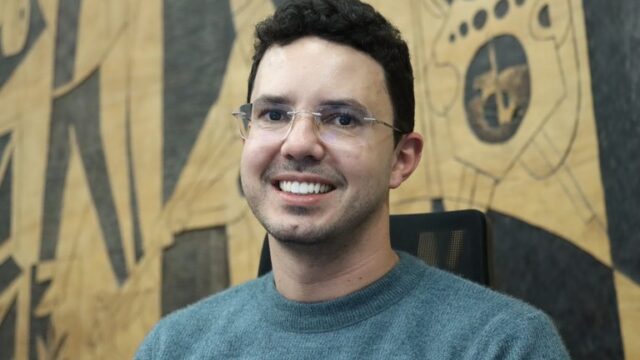 Retrato de homem sorrindo com óculos e suéter azul em frente a painel de madeira