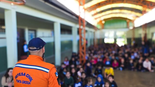 Agente da Defesa Civil de costas, vestindo uniforme laranja, palestrando para dezenas de alunos sentados em um pátio escol…