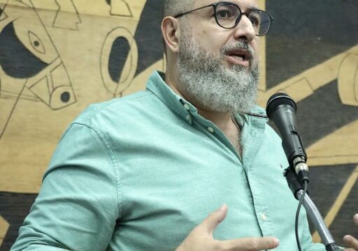 Homem com barba e óculos veste camisa verde e fala ao microfone em tribuna