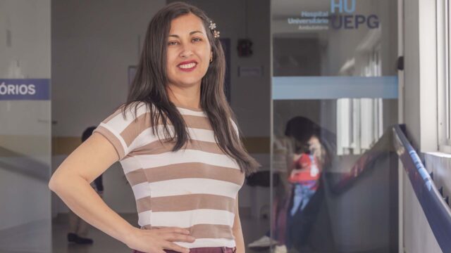 Mulher com blusa listrada e cabelo solto posa sorrindo em corredor do hospital universitário